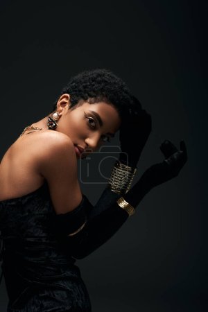 Trendy junge afrikanisch-amerikanische Frau in goldenen Accessoires, Handschuhen und Kleid, die Haare berühren und isoliert in die Kamera schauen auf schwarz, High Fashion und Abendlook