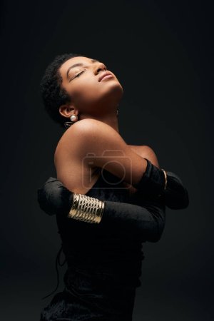 Foto de Mujer afroamericana de pelo corto con maquillaje de noche que usa guantes, vestido y accesorios dorados mientras posa aislada en negro, alta moda y look de noche, femenino - Imagen libre de derechos