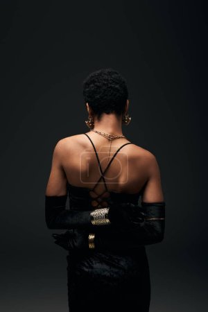 Vista posterior de la mujer afroamericana elegante y de pelo corto en guantes, vestido y accesorios modernos de pie aislado en negro, alta moda y aspecto de noche, femenino