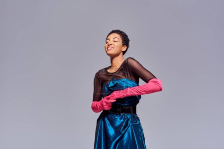 Fröhliche und stilvolle junge afrikanisch-amerikanische Frau in rosa Handschuhen und Cocktailkleid, die mit geschlossenen Augen auf grauem, modernem Modekonzept der Generation z steht