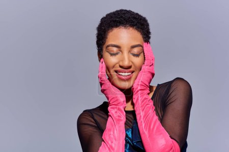 Fröhliche afrikanisch-amerikanische Frau mit kühnem Make-up und rosa Handschuhen, die Augen schließen und das Gesicht berühren, während sie isoliert auf einem grauen, modernen Modekonzept der Generation z steht