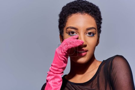 Portrait of fashionable african american woman with bold makeup and pink glove touching face and smiling isolated on grey, modern generation z fashion concept