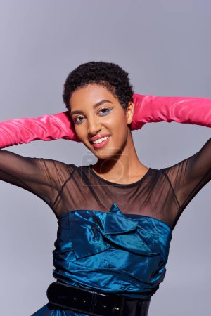 Smiling and fashionable african american woman with bold makeup wearing pink gloves and cocktail dress and touching head isolated on grey, modern generation z fashion concept