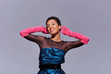 Joyful and short haired african american woman with bold makeup wearing pink gloves and cocktail dress while standing and posing isolated on grey, modern generation z fashion concept