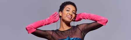 Smiling and trendy african american woman with bold makeup wearing pink gloves and posing while standing isolated on grey, modern generation z fashion concept, banner 