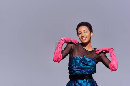 Trendy young african american woman in pink gloves and cocktail dress touching shoulders and smiling isolated on grey, modern generation z fashion concept