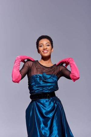 Positive afrikanisch-amerikanische Frau in Cocktailkleid und rosa Handschuhen, die die Schultern berührt und vereinzelt in die Kamera blickt, auf grauem, modernem Modekonzept der Generation Z