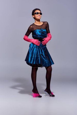 Volle Länge der fröhlichen und stilvollen afrikanisch-amerikanischen Frau in Sonnenbrille, Cocktailkleid und High Heels mit Federn posiert auf grauem Hintergrund, modernes Modekonzept der Generation Z