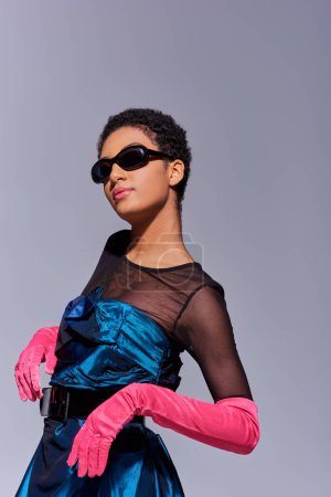 Confident young african american woman in sunglasses, pink gloves and cocktail dress posing and standing isolated on grey, modern generation z fashion concept