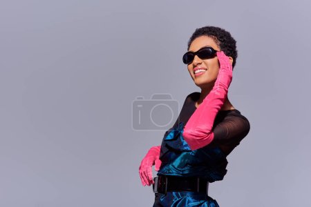 Fröhliche afrikanisch-amerikanische Frau in Cocktailkleid und rosa Handschuhen, die Sonnenbrille berührt und isoliert auf grauem, modernem Modekonzept der Generation Z steht