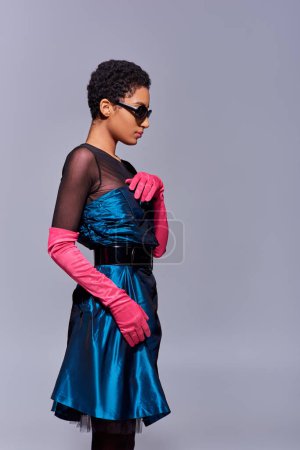 Trendy young african american woman in cocktail dress, sunglasses and pink gloves touching bow while standing isolated on grey, modern generation z fashion concept