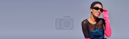 Foto de Joven mujer afroamericana alegre en gafas de sol con estilo, guante rosa y vestido de cóctel posando mientras está de pie aislado en gris, concepto de moda z generación moderna, bandera - Imagen libre de derechos