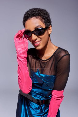 Porträt eines jungen, lächelnden und kurzhaarigen afrikanisch-amerikanischen Modells in rosa Handschuhen und Cocktailkleid, das Sonnenbrillen auf grauem, modernem Modekonzept der Generation Z berührt