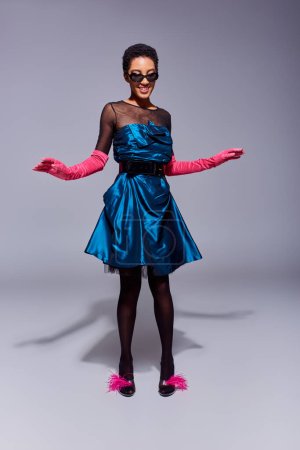 Volle Länge der fröhlichen afrikanisch-amerikanischen Frau mit Sonnenbrille, Cocktailkleid und rosa Handschuhen, die in Federschuhen posiert, während sie auf grauem Hintergrund steht, modernes Modekonzept der Generation Z