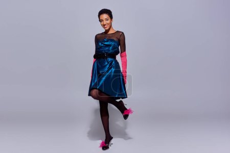 Longitud completa de la mujer afroamericana positiva en guantes de color rosa, vestido de cóctel y zapatos emplumados posando y de pie sobre fondo gris, concepto de moda z generación moderna
