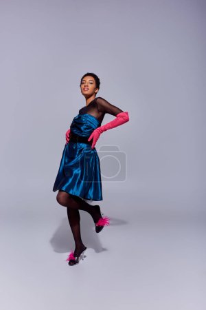Femme afro-américaine tendance en robe de cocktail, gants roses et talons avec des plumes touchant les hanches et souriant tout en se tenant debout sur fond gris, concept de mode moderne génération z