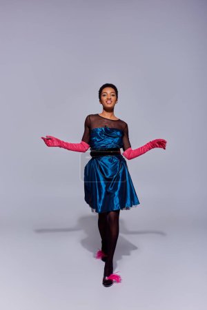 Longitud completa de alegre modelo afroamericano en guantes de color rosa, vestido de cóctel y tacones con plumas caminando y mirando a la cámara sobre fondo gris, concepto de moda z generación moderna