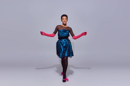 Mujer afroamericana sonriente y de pelo corto en vestido de cóctel, guantes rosas y tacones con plumas caminando sobre fondo gris, concepto de moda z generación moderna