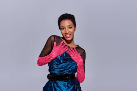 Portrait of positive and stylish african american model with bold makeup in pink gloves and cocktail dress looking at camera isolated on grey, modern generation z fashion concept