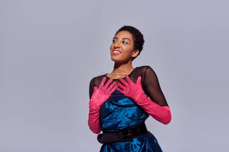 Positives afrikanisch-amerikanisches Model mit kurzen Haaren posiert in rosa Handschuhen und Cocktailkleid, während sie mit Händen nahe der Brust isoliert auf grauem, modernem Modekonzept der Generation Z steht