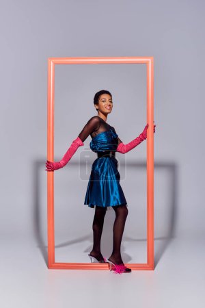 joyful african american model in evening dress, pink gloves and heels with feathers looking at camera near frame on grey background, modern generation z fashion concept