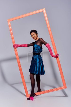 modèle afro-américain positif et confiant en robe de cocktail, gants roses et talons à plumes tenant cadre et debout sur fond gris, génération z moderne, concept de mode