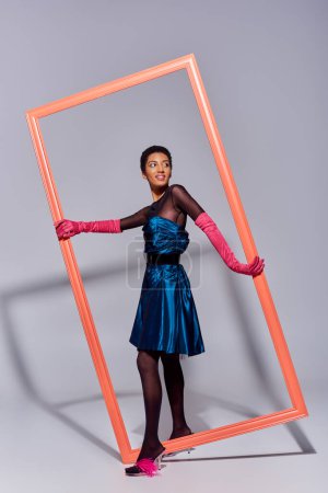 Young and smiling african american woman in pink gloves, cocktail dress and heels with feathers looking away near frame on grey background, modern, generation z fashion concept