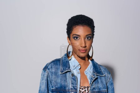 Glamorous african american woman with bold makeup and golden earrings wearing denim jacket and looking at camera while standing on grey background, denim fashion concept