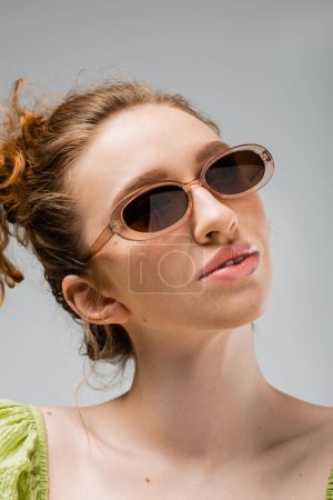 Porträt einer jungen rothaarigen und sommersprossigen Frau in grüner Bluse und trendiger Sonnenbrille, die isoliert auf grauem, trendigem Sonnenschutzkonzept und Modemodel posiert 