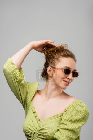 Lächelnde junge rothaarige Frau in sommergrüner Bluse und Sonnenbrille, die die Haare berührt, während sie isoliert auf grauem Hintergrund steht, trendiges Sonnenschutzkonzept, Modemodel