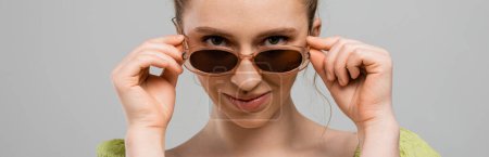 Portrait of modern young woman with natural makeup touching stylish sunglasses and looking at camera isolated on grey background, trendy sun protection concept, banner, fashion model 