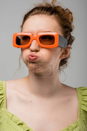 Portrait of young redhead woman in sunglasses and green blouse grimacing and pouting lips while standing isolated on grey background, trendy sun protection concept, fashion model 