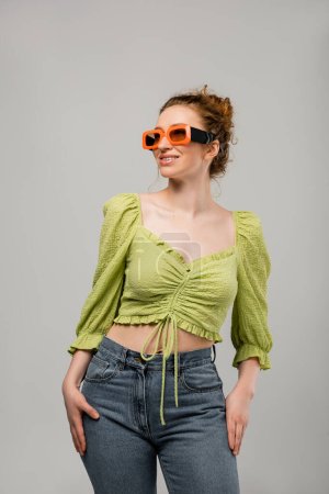 Fröhliche rothaarige Frau in Jeans, grüner Bluse und Sonnenbrille posiert und schaut weg, während sie isoliert auf grauem Hintergrund steht, trendiges Sonnenschutzkonzept, Modemodel 