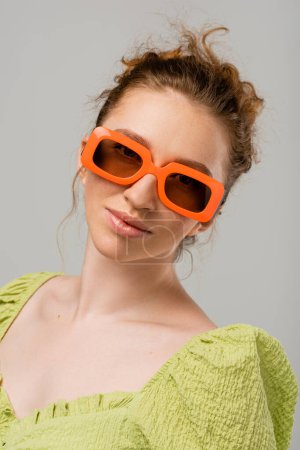 Retrato de mujer pelirroja joven en blusa verde y gafas de sol modernas mirando a la cámara mientras está de pie aislado sobre fondo gris, concepto de protección solar de moda, modelo de moda 