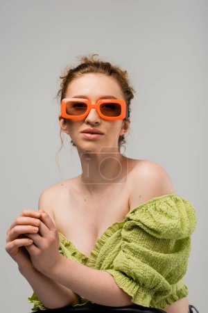 Retrato de mujer de pelo rojo joven con estilo en gafas de sol y blusa verde con hombros desnudos posando mientras está de pie aislado sobre fondo gris, concepto de protección solar de moda, modelo de moda 