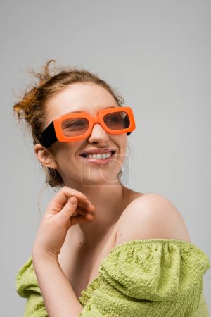 Portrait of cheerful young red head woman in modern sunglasses and green blouse with naked shoulders looking away and posing isolated on grey background, trendy sun protection concept, fashion model 