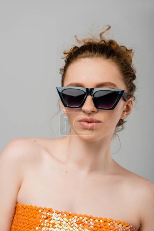 Retrato de mujer de moda y pelirroja en gafas de sol y top naranja con lentejuelas y hombros desnudos de pie aislados sobre fondo gris, concepto de protección solar de moda, modelo de moda 
