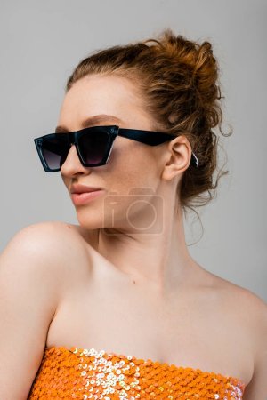 Fashionable young redhead woman in sunglasses and top with orange sequins and naked shoulders posing isolated on grey background, trendy sun protection concept, fashion model 