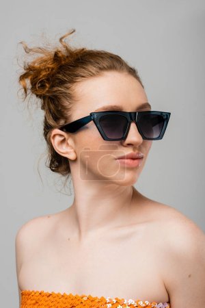 Porträt einer jungen rothaarigen und sommersprossigen Frau mit Sonnenbrille und Top mit Pailletten, die auf grauem Hintergrund isoliert dasteht, trendiges Sonnenschutzkonzept, Modemodel 