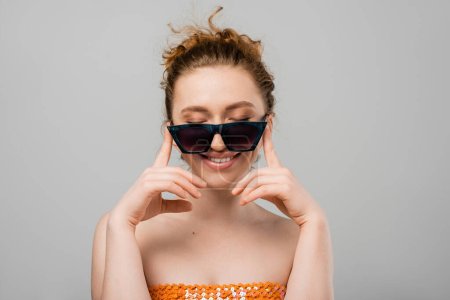Fröhliche junge rothaarige Frau mit natürlichem Make-up in Sonnenbrille und orangefarbenem Top mit Pailletten, die Augen schließen, während sie isoliert auf grauem Hintergrund steht, trendiges Sonnenschutzkonzept, Modemodel 