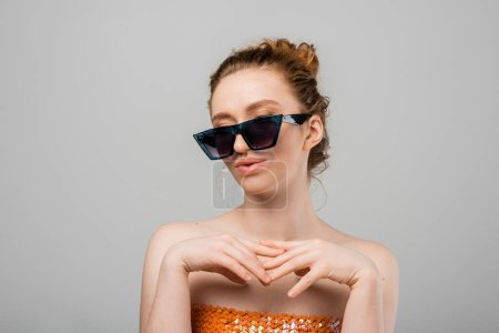 Jeune femme rousse en haut avec paillettes orange et épaules nues posant dans des lunettes de soleil et debout isolé sur fond gris, concept de protection solaire à la mode, mannequin 