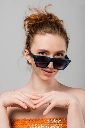Retrato de joven modelo de pelo rojo con maquillaje natural y hombros desnudos mirando a la cámara y posando en gafas de sol aisladas sobre fondo gris, concepto de protección solar de moda, modelo de moda 