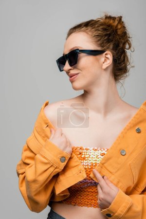 Joyeux rousse femme avec maquillage naturel dans les lunettes de soleil, veste en denim orange et haut avec paillettes posant et debout isolé sur fond gris, concept de protection solaire à la mode, modèle de mode 