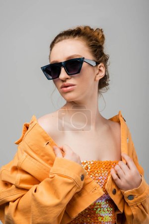 Fashionable redhead woman with natural makeup in sunglasses and top with sequins touching orange jacket while standing isolated on grey background, trendy sun protection concept, fashion model 