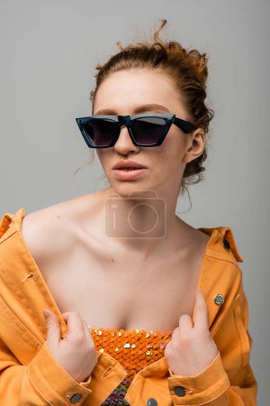 Young and redhead woman in sunglasses and top with orange sequins touching orange denim jacket and posing isolated on grey background, trendy sun protection concept, fashion model 