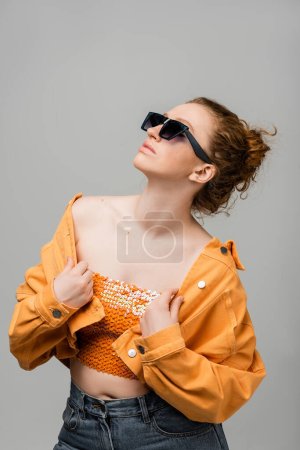 Elegante mujer pelirroja joven en gafas de sol y la parte superior con lentejuelas tocando chaqueta de mezclilla naranja y de pie y posando aislado sobre fondo gris, concepto de protección solar de moda, modelo de moda 