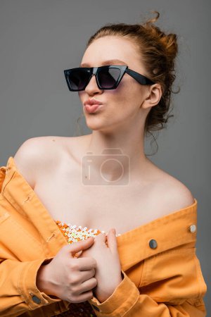 Porträt einer jungen rothaarigen Frau mit Sonnenbrille, Top mit Pailletten und orangefarbener Jeansjacke, schmollende Lippen und isoliertes Posieren auf grauem Hintergrund, trendiges Sonnenschutzkonzept, Modemodel 