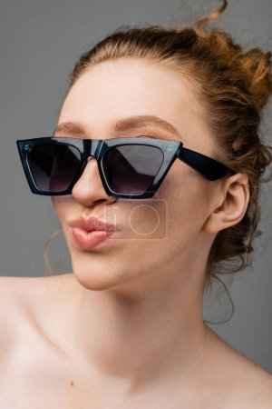 Nahaufnahme der jungen rothaarigen und sommersprossigen Frau in stylischer Sonnenbrille mit nackten Schultern schmollenden Lippen und isoliert auf grauem Hintergrund stehend, trendiges Sonnenschutzkonzept, Modemodel 