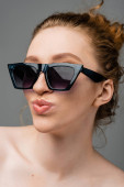Close up view of young redhead and freckled woman in stylish sunglasses with naked shoulders pouting lips and standing isolated on grey background, trendy sun protection concept, fashion model  magic mug #660903746