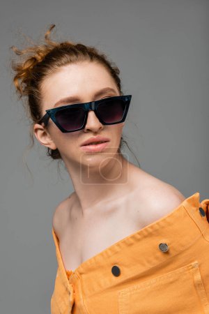 Portrait de mannequin roux et taches de rousseur en lunettes de soleil et veste en denim orange posant et debout isolé sur fond gris, concept de protection solaire tendance, mannequin 